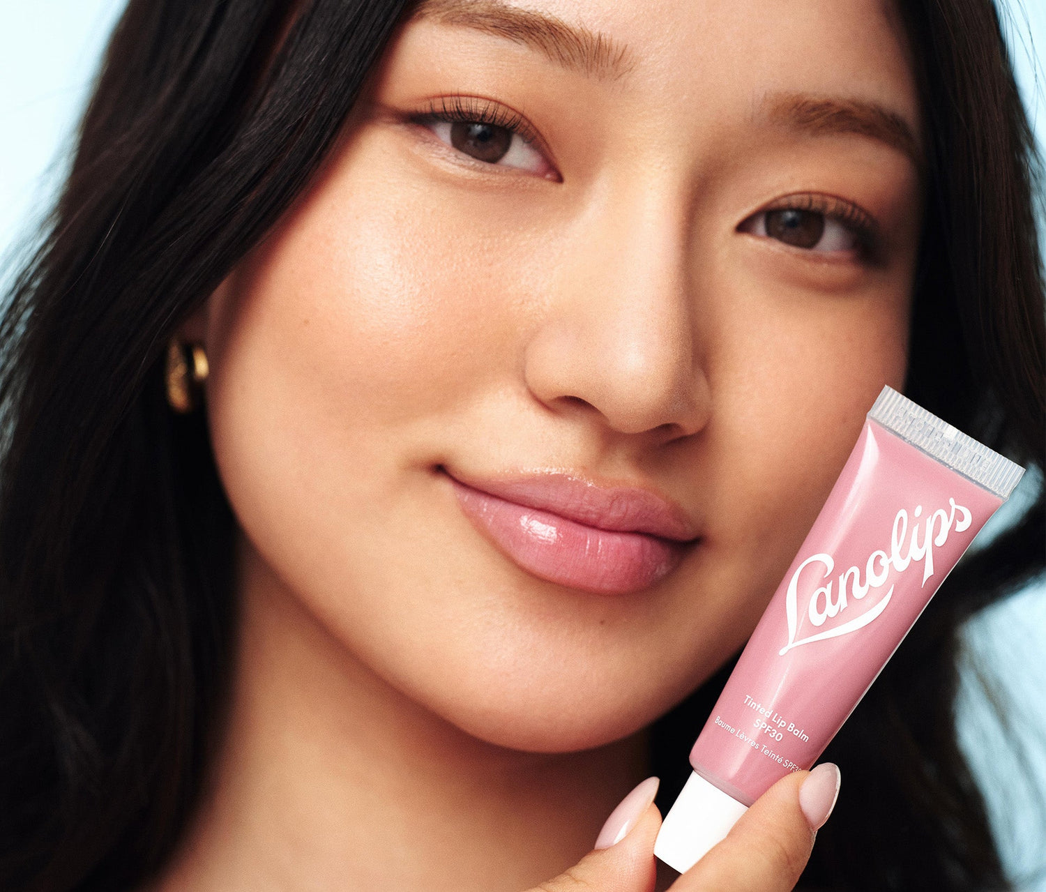 A model wears the Lanolips Tinted Lip Balm SPF30 Rhubarb on her lips. She also holds the tube of the Tinted Lip Balm in Rhubarb, a fruity pink shade designed to enhance natural lip colour. This cult-favourite formula is made with lanolin, vitamin e, shea butter and SPF30 for a long-lasting, non-sticky finish. With the right amount of pigment and high-gloss finish, it's the perfect everyday lip balm. The SPF30 helps shield lips from sun damage.