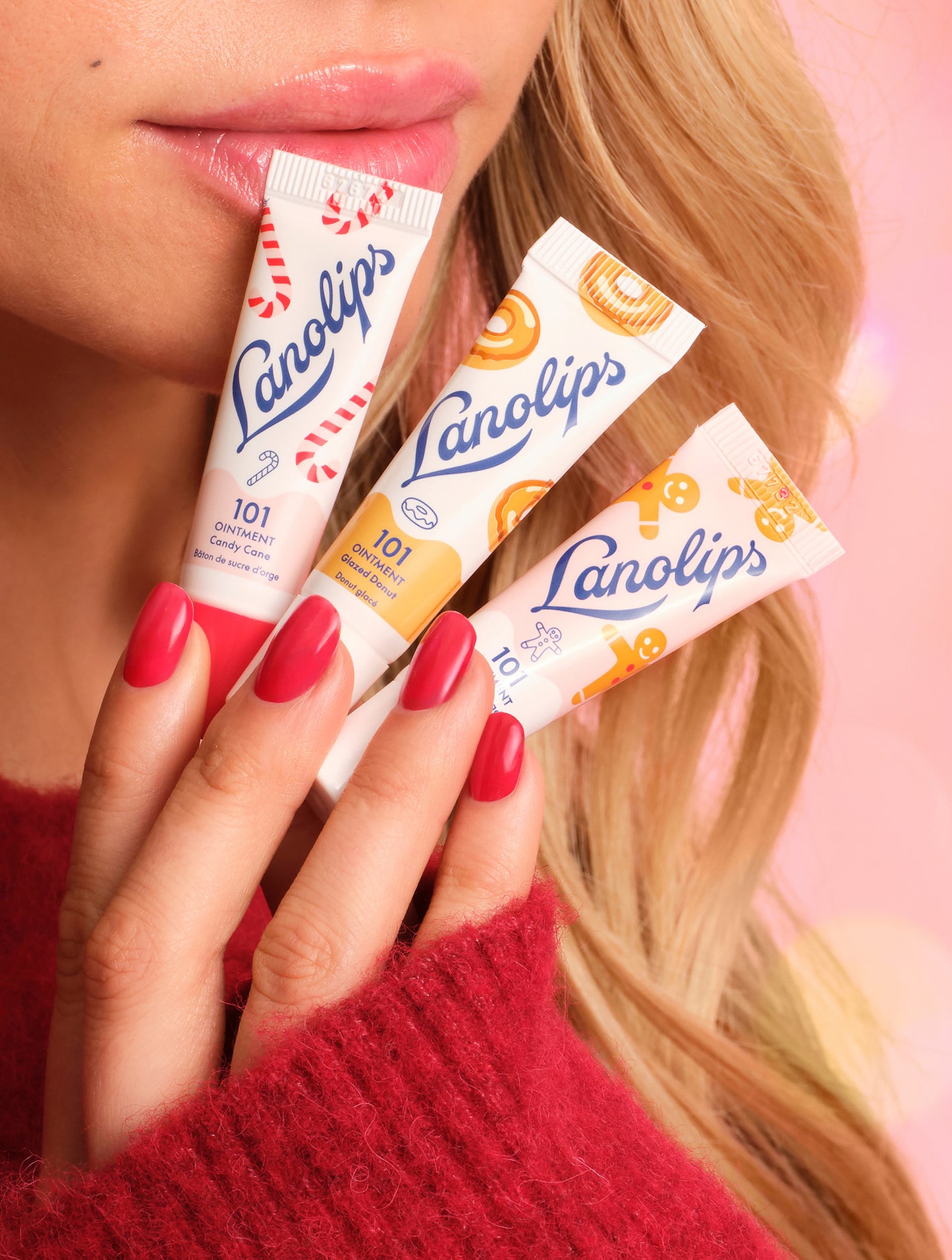 Close-up of model holding Lanolips Holiday Mini Bites lip balm trio in Candy Cane, Gingerbread and Glazed Donut flavours, with glossy lips and red nails.