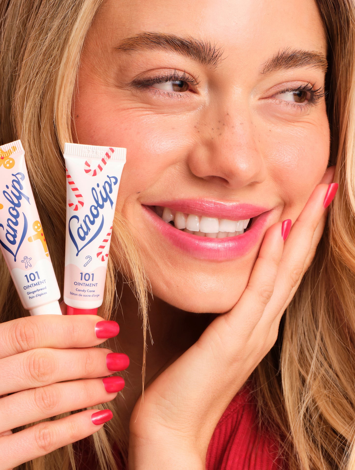 Model smiling while holding Lanolips 101 Ointment Multi-Balm Gingerbread & Candy Cane, made with lanolin, vitamin E and natural flavours, from the limited-edition Holiday Gingerbread House collection.